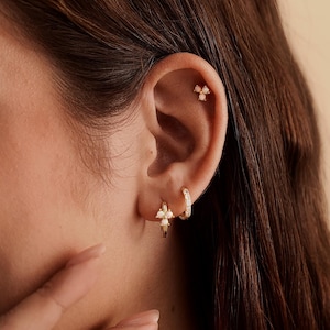 May include: A gold hoop earring featuring a three-stone opal cluster charm, a small gold hoop earring with a textured surface, and a gold stud earring with a three-stone opal cluster, all worn in the earlobe and cartilage piercings.
