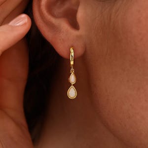 Könnte beinhalten: Ein goldener Creolen-Ohrring mit zwei hängenden, tropfenförmigen weißen Opalsteinen. Der Ohrring ist am Ohrläppchen befestigt und zeigt ein poliertes Gold-Finish sowie schillernde Opal-Akzente in einem zarten, vertikalen Hängedesign.
