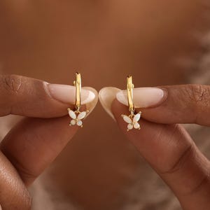 May include: A pair of gold hoop earrings, each featuring a small, white, iridescent butterfly-shaped charm. The earrings are polished and metallic, with the butterfly charms suspended from the bottom of each circular hoop.