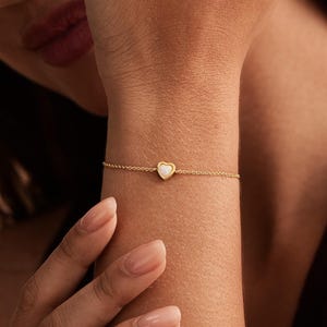 May include: A delicate gold chain bracelet featuring a small, centered heart-shaped charm with an iridescent opal inlay. The thin, polished metal chain secures around the wrist, highlighting the subtle shimmer of the white stone set in gold.
