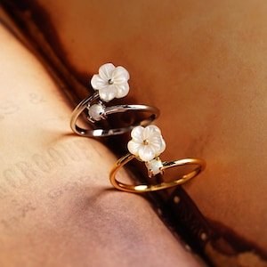 May include: Two open-style rings, one in silver-tone and one in gold-tone metal. Each ring features a carved white floral motif and a small, round, iridescent stone set on the opposite end of the open band, creating a delicate, layered appearance.