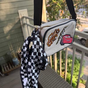 May include: A rectangular beige crossbody bag with a black adjustable strap and a gold zip. The front features embroidered patches of a brown tow truck, a yellow lightning bolt, and a red race car. A black and white chequered scarf is attached.