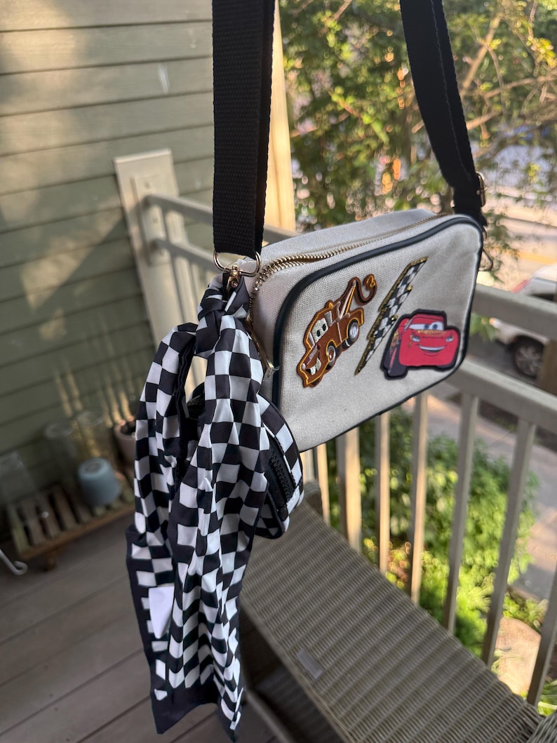 May include: A rectangular beige crossbody bag with a black adjustable strap and a gold zip. The front features embroidered patches of a brown tow truck, a yellow lightning bolt, and a red race car. A black and white chequered scarf is attached.