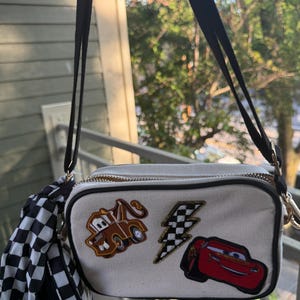 May include: A rectangular off-white crossbody bag featuring embroidered patches of a brown tow truck, a red race car, and a black and white checkered lightning bolt. The bag includes a black adjustable strap and a black and white checkered accessory.
