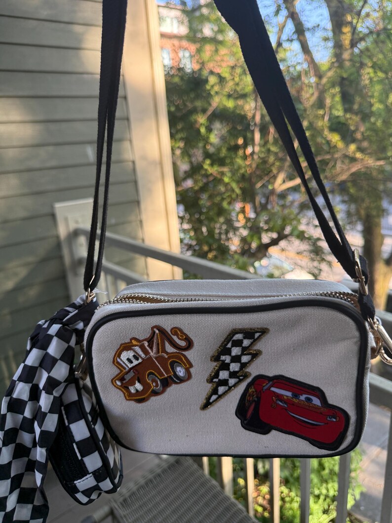 May include: A rectangular off-white crossbody bag featuring embroidered patches of a brown tow truck, a red race car, and a black and white checkered lightning bolt. The bag includes a black adjustable strap and a black and white checkered accessory.