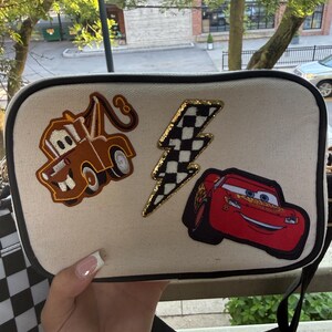 May include: A rectangular off-white canvas crossbody bag with black trim. The front features three embroidered patches: a brown tow truck, a black and white chequered lightning bolt with a gold border, and a red race car with the number 95.
