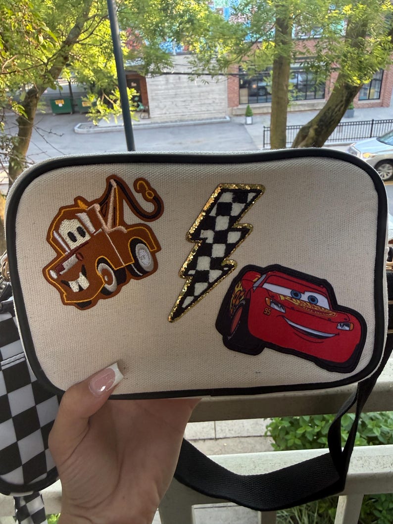 May include: A rectangular off-white canvas crossbody bag with black trim. The front features three embroidered patches: a brown tow truck, a black and white chequered lightning bolt with a gold border, and a red race car with the number 95.