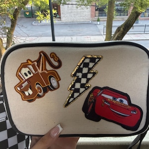 May include: A rectangular off-white canvas crossbody bag with black trim. The front features three embroidered patches: a brown tow truck, a black and white chequered lightning bolt with gold glitter edges, and a red race car with the number 95.