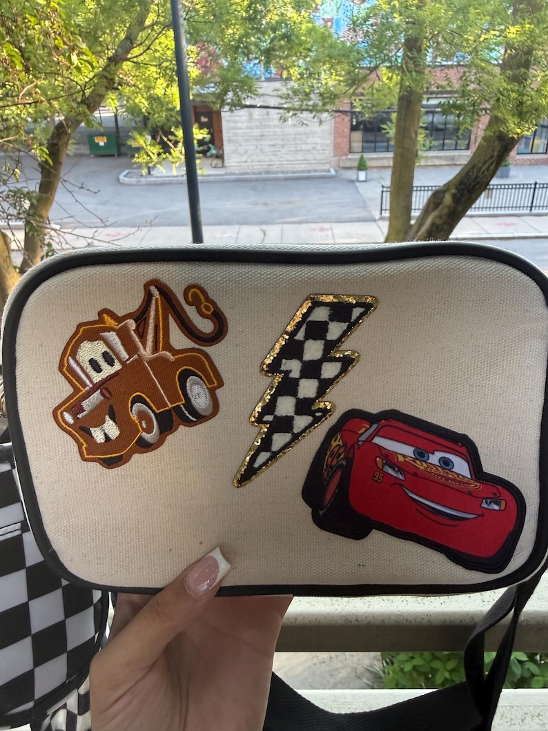 May include: A rectangular off-white canvas crossbody bag with black trim. The front features three embroidered patches: a brown tow truck, a black and white chequered lightning bolt with gold glitter edges, and a red race car with the number 95.