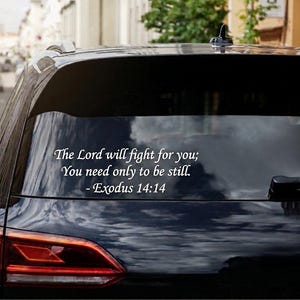 Può includere: Un adesivo in vinile bianco applicato sul lunotto posteriore di un veicolo nero. L'adesivo riporta il testo: The Lord will fight for you; You need only to be still. - Exodus 14:14, scritto in un carattere serif bianco sulla superficie scura.
