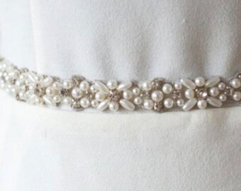 May include: A white beaded sash with pearl and rhinestone embellishments. The sash is a delicate and elegant accessory for a wedding or special occasion.