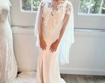 May include: A bride wearing a white wedding dress with a sheer, beaded cape. The dress is a simple, elegant design with a high slit. The cape features floral embroidery and pearl accents. The bride is smiling in front of a window.