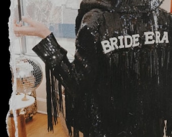 May include: A black sequined jacket with the text "BRIDE ERA" in white. The jacket has long black fringe and is worn by a person with blonde hair. A disco ball and small table are visible in the background.