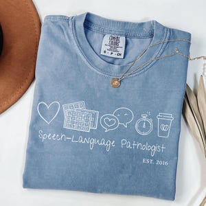 May include: A blue crew neck t-shirt featuring white line art graphics of a heart, communication boards, speech bubbles, a stopwatch, and a coffee cup. The text reads Speech-Language Pathologist and EST. 2016. A gold necklace with a round pendant rests on top.