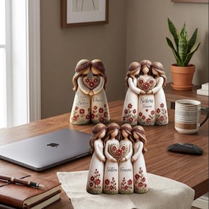 May include: Three decorative figurines featuring stylised figures with long brown hair, wearing cream-coloured dresses with red floral patterns. One figurine depicts two figures holding a heart, while the others show three and four figures holding a heart.