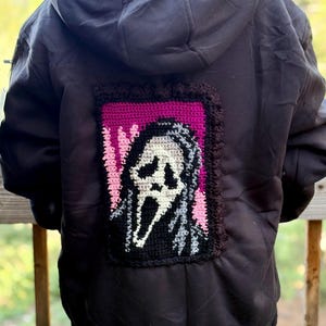 May include: A black hooded jacket featuring a rectangular crochet patch on the back. The patch depicts a white ghost-like mask with black eye and mouth openings against a vibrant pink and black background, framed by a dark brown crocheted border.