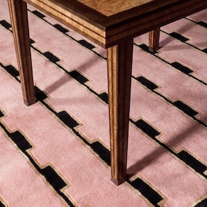 Op de afbeelding: Een houten tafel met een gepolijst blad van lichtbruin wortelhout en donkerbruine randen, ondersteund door vier rechte, verticaal gestreepte houten poten. De tafel staat op een zacht, getextureerd tapijt met een herhalend geometrisch patroon.