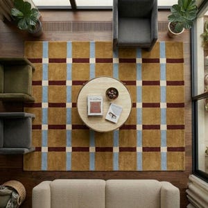 May include: A large rectangular area rug featuring a grid pattern of mustard yellow, light blue, and dark maroon stripes with cream-coloured intersections. The rug is positioned under a round light-coloured coffee table with two books on top.