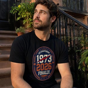 May include: A black short-sleeved t-shirt featuring a graphic print with the text "LAST CHAMPIONSHIP: 1973 2026 FINALLY." The design includes a red lightning bolt, basketball icons, and a laurel wreath border in an oval shape.