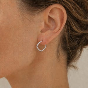 Op de afbeelding: Een kleine, zilverkleurige vierkante oorring met een gepolijste afwerking. De oorbel bestaat uit een dunne, doorlopende metalen band in de vorm van een afgerond vierkant, ontworpen om in de oorlel te dragen met een eenvoudig sluitmechanisme.