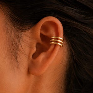 Peut inclure: Un ear cuff doré composé de trois anneaux parallèles et lisses qui entourent le cartilage externe de l'oreille. Le métal présente une finition polie et réfléchissante qui capte la lumière, offrant un aspect net et uniforme sur le cartilage.