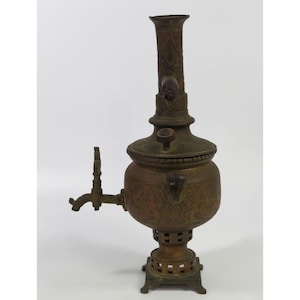 May include: A tall, antique-style brass samovar featuring an ornate, engraved floral pattern across its rounded body and chimney. The vessel includes a side-mounted spigot, a removable lid with a small handle, and a square, perforated base stand.