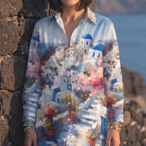 May include: A button-down shirt with a vibrant print depicting a coastal village with white buildings, blue domes, and colorful flowers. The shirt's color palette includes shades of blue, white, pink, and green, creating a summery aesthetic.