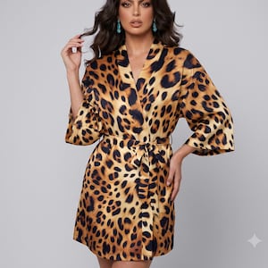 May include: A short robe featuring a bold leopard print pattern in shades of tan, brown, and black. The garment has a shawl collar, wide three-quarter length sleeves, and a matching fabric tie belt that secures at the waist for a wrap closure.