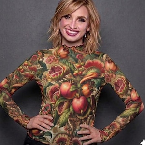 Puede incluir: Una camiseta de manga larga con cuello alto, con un estampado botánico vibrante de manzanas, granadas y otras frutas y follaje en tonos rojos, verdes y marrones. La camiseta tiene una silueta ajustada y un ribete con volantes.