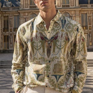 May include: A cream-coloured button-down shirt with a detailed pattern featuring birds, architectural elements, and foliage in shades of green, red, and blue. The shirt is worn with light-coloured trousers.