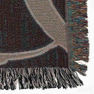 May include: A woven throw blanket with a geometric pattern in shades of brown, beige, and black. The blanket has a fringe border.