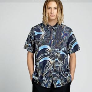 Könnte beinhalten: Kurzärmeliges Hemd mit einem einzigartigen, abstrakten Muster in Blau-, Schwarz- und schillernden Farbtönen. Das Hemd hat einen klassischen Kragen und eine lockere Passform. Das Design zeigt ein wirbelndes, flüssigkeitsartiges Motiv.