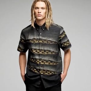 Puede incluir: Camisa negra de manga corta con un patrón único de líneas onduladas horizontales doradas y grises. La camisa tiene un cuello clásico y se usa con pantalones negros. El diseño de la camisa es llamativo y moderno.