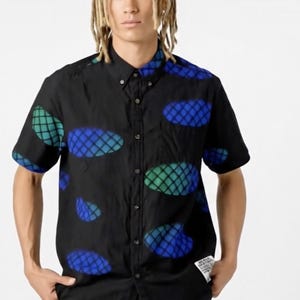 Puede incluir: Camisa negra de manga corta con un estampado de formas ovaladas azules y verdes. La camisa tiene un cuello clásico y un pequeño bolsillo. El diseño se repite en toda la camisa. La camisa está hecha de tela ligera.