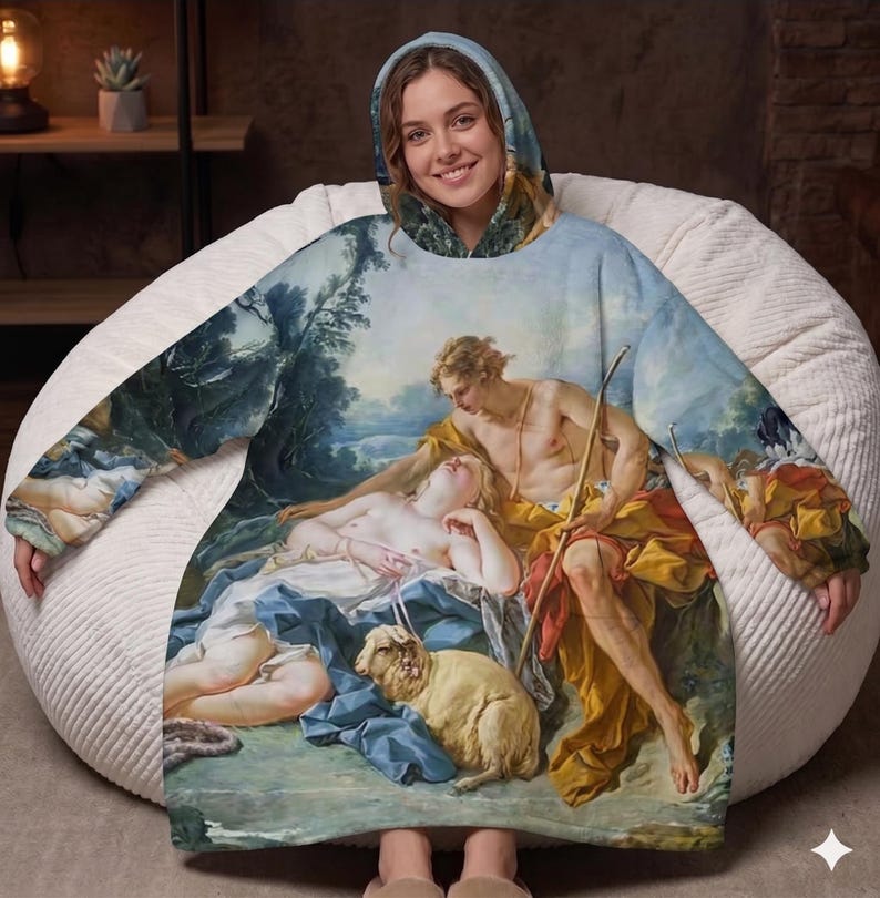 May include: A hooded blanket featuring a classical painting of a reclining figure with two other figures and a sheep. The blanket has a blue, yellow, and white color scheme, with a hood and long sleeves. The person wearing the blanket is smiling.
