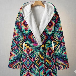 Op de afbeelding: Een badjas met capuchon met een levendig geometrisch patroon in tinten van turquoise, paars, geel en roze. De badjas heeft een witte, zachte binnenkant en een bijpassende riem. De badjas hangt aan een houten kleerhanger.