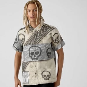 Può includere: Camicia a maniche corte con motivo a teschio. La camicia ha una base color crema con grafica a teschio grigia e un pannello grigio sul petto. Il colletto e le maniche sono risvoltati. Una piccola etichetta è visibile in basso.