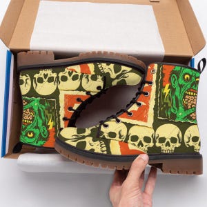 Puede incluir: Botas militares con suelas marrones y cordones negros. Las botas presentan un patrón repetido de calaveras y gráficos de zombis en verde, amarillo y naranja. Las botas están en una caja de cartón.