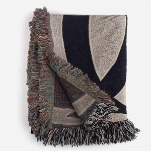 May include: A woven throw blanket with a geometric pattern in shades of beige and navy blue. The blanket has a fringe border.