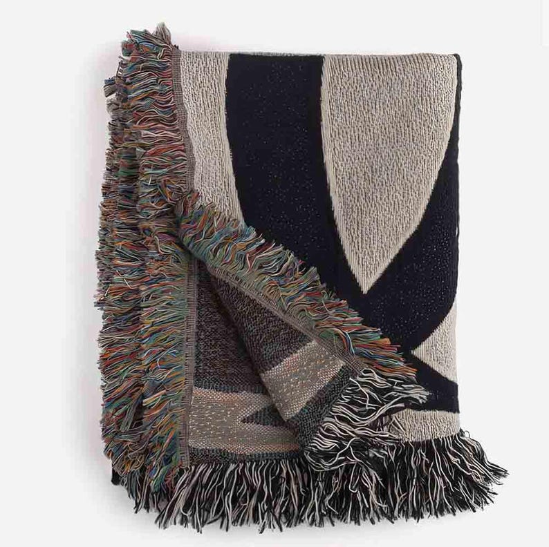 May include: A woven throw blanket with a geometric pattern in shades of beige and navy blue. The blanket has a fringe border.