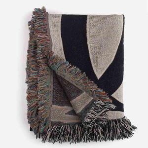 May include: A woven throw blanket with a geometric pattern in shades of beige and navy blue. The blanket has a fringe border.