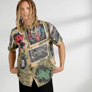 Può includere: Camicia a maniche corte con bottoni e motivo mimetico. La camicia presenta varie toppe grafiche, tra cui un personaggio diavolo, una faccia di mostro e un boombox. La camicia è indossata da una persona con i dreadlocks.
