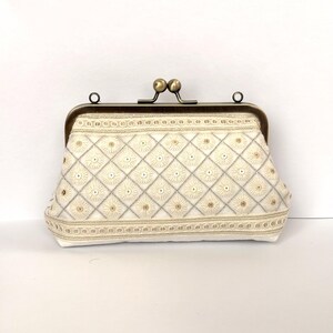 May include: A cream-colored clutch purse featuring a diamond-patterned embroidery with small circular floral motifs. The bag has a metallic bronze-toned kiss-lock frame closure at the top and decorative horizontal trim along the bottom edge.