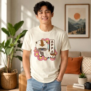 May include: A cream-colored short-sleeve t-shirt featuring a graphic of the character Appa from Avatar: The Last Airbender. The design includes a brown arrow on the creature's head, pink flowers in the background, and the text APPA vertically.