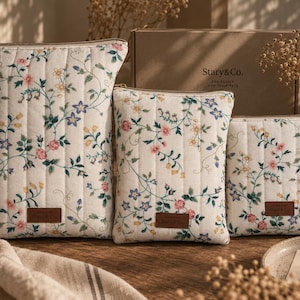 May include: Three rectangular quilted pouches in varying sizes, featuring an off-white fabric with a delicate floral pattern of pink, blue, and yellow flowers. Each pouch has a brown leather-like label with the text Stary & Co. and a zipper.
