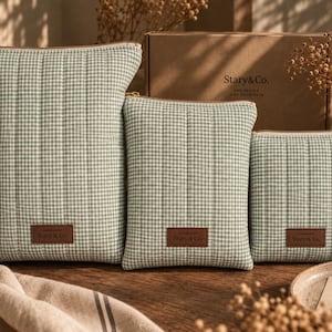 May include: Three rectangular quilted pouches in a sage green and white gingham pattern, each featuring a gold zipper and a brown leather-like rectangular patch embossed with the text Stary & Co. The pouches are arranged in descending size order.