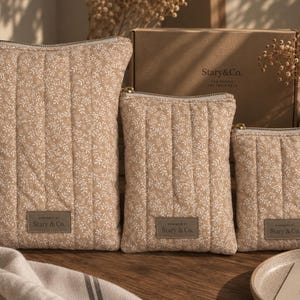 May include: Three quilted pouches in varying sizes, featuring a tan fabric with a white floral pattern. Each pouch has a gold-tone zipper and a rectangular grey label with the text Stary & Co. stitched onto the lower front section.