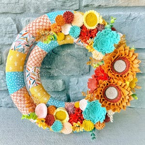 May include: A circular wreath featuring a fabric-wrapped frame with alternating patterns of blue dots, orange geometric shapes, and yellow floral prints. The wreath is decorated with felt flowers in shades of turquoise, mustard yellow, and orange.