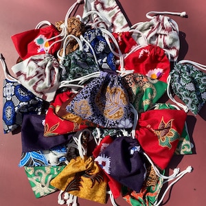 May include: A collection of small fabric drawstring pouches featuring a variety of colorful patterns, including floral, geometric, and batik-style prints in shades of red, blue, green, yellow, and purple, each with a white cord closure.