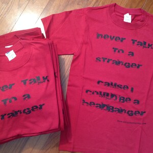 May include: Two red t-shirts with black text. The text on the top shirt reads "never talk to a stranger cause I could be a heartbreaker". The text on the bottom shirt reads "never talk to a stranger".